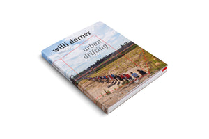 A photograph of the book urban drifting by Willi Dorner, displayed at an angle on a white background. The cover features a long line of people in colorful clothing standing on a dusty hillside under a blue, cloud-filled sky.