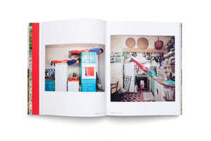 An open photobook on a white background. The left page shows a photo of a man and a woman in red pants and blue shirts planking on top of kitchen appliances. The right page shows a photo of a person in a similar outfit doing a handstand on the counter of a cluttered kitchen, their body arched backward.
