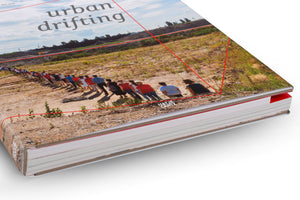 An angled, close-up photograph of a hardcover book titled urban drifting. The cover image shows a long line of people from behind, standing in single file on a dusty, barren hillside under a cloudy sky. Thin red lines form a graphic frame over the photo. The book rests on a white surface, showing the thickness of its pages which have a red edge.