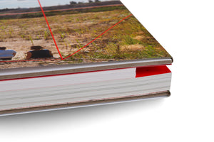 A close-up, angled shot of a thick, hardcover book on a white background. The cover shows a photo of two people sitting in a grassy field, with red lines overlaid. The edge of the book is visible, showing the stacked white pages and a red-colored section with a page number tab.