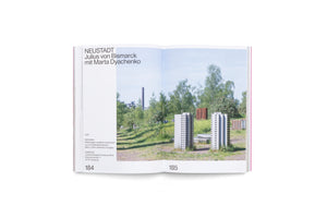 An open book showing a two-page spread. The left page, 184, contains German text for an artwork titled NEUSTADT. The right page, 185, is a photograph of an outdoor installation featuring miniature concrete models of tower blocks arranged in a grassy field with trees and a factory in the background.