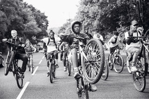 A dynamic black and white photograph of a group of diverse people riding bicycles down a tree-lined street. In the foreground, a young man in a hoodie stares intently at the camera while performing a wheelie, his front wheel high in the air. Others in the group ride alongside him, also doing wheelies.