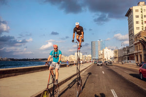 A wide, eye-level shot of two men riding very tall bicycles along a sunny seaside road in Havana, Cuba. One man rides on the sidewalk next to the sea, while the other stands on the pedals of an even taller bike on the road, with classic cars and city buildings in the background.