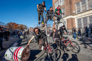 A group of people joyfully rides a massive, multi-level, custom-built bicycle down a city street during an event. Three men are on an upper platform, while on the ground level, a man steers while dragging a large plastic barrel, and a woman sits on a swing attached to the bike's frame.