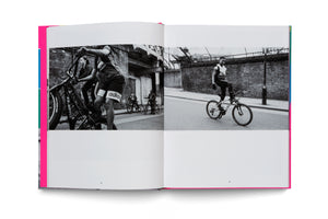 An open book with a bright pink spine shows a two-page spread of black and white street photography. On the left, several young people are gathered on their bikes in an urban setting. On the right, a smiling young man stands on his bicycle's pedals in the middle of a street.