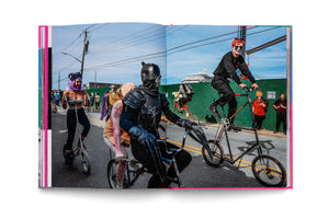 A two-page photo spread in an open book shows a vibrant street parade on a sunny day. People in eclectic costumes ride customized tall bikes and scooters. In the foreground, a person in a black armored suit rides a bike with a passenger. To their right, a person with red hair and dramatic face paint rides a very tall bicycle. Other costumed participants are visible in the background, with a large cruise ship seen over a green wall.