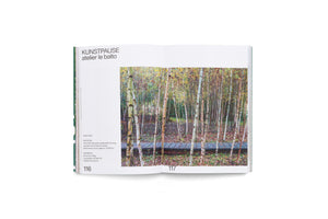 An open book on a white surface. The left page, numbered 116, has text describing the KUNSTPAUSE project by atelier le balto. The right page, 117, features a full-page photo of a wooden boardwalk passing through a dense forest of slender birch trees during autumn.
