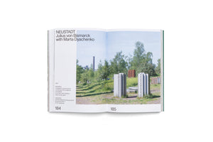 An open book showing a two-page spread about an art installation. The left page, 184, contains text including the title NEUSTADT and artist Julius von Bismarck. The right page, 185, displays a photograph of the installation: miniature concrete sculptures of modernist buildings situated in a grassy field with trees and an industrial smokestack in the background.