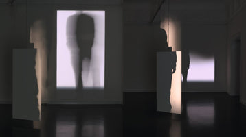 A dimly lit art installation in a gallery with a dark, reflective floor. Blurry projections of human figures appear on the walls, while tall, white rectangular panels hang from the ceiling, casting distinct, person-shaped shadows. The scene creates a mysterious interplay of light and form.