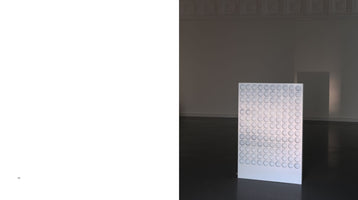An art catalog spread with a blank left page numbered 142. The right page shows a minimalist gallery with a freestanding white rectangular panel on a dark floor. The panel's surface is covered in a uniform grid of circular, concave indentations.