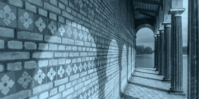 A low-angle, blue-toned photograph of a long, covered walkway beside a river. On the left is a brick wall with decorative floral tiles, onto which arched shadows are cast. On the right, a row of fluted columns opens up to a view of the water and the opposite bank.