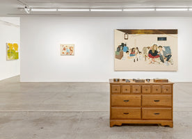 An installation view of an art gallery with white walls and a concrete floor. In the foreground is a wooden dresser with small sculptures on top. Behind it hangs a large painting of a group of people in a room. Two smaller, more abstract paintings are visible on the walls in the background.