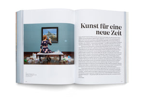 An open book viewed from above. The left page shows a photograph of a woman sitting on a museum bench, her back to the camera, surrounded by a large pile of empty plastic bottles. She looks at a framed painting of a dramatic, rocky landscape. The right page contains a block of German text under the large headline, Kunst für eine neue Zeit.
