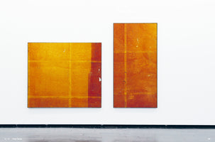 An eye-level view of two large, abstract rectangular paintings hanging side-by-side on a white gallery wall over a dark floor. The painting on the left is horizontal, rendered in golden yellow and orange with a faint grid and a thick red stripe on the right. The painting on the right is vertical, a deeper reddish-orange, also with a subtle grid pattern. The image appears as a two-page spread in a book or catalog.