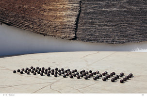 A wide shot of an outdoor art installation featuring dozens of dark, round objects arranged in loose rows on a light-colored, cracked stone patio. In the background, a stark white wall is overshadowed by the uneven edge of a thick, two-toned thatched roof.