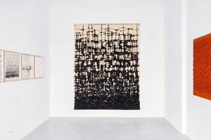 An interior view of a modern art gallery with white walls and a grey floor, featuring a large, central abstract artwork. The piece is a vertical canvas with a dense, grid-like pattern of thick black lines over a light background. To the left, a series of smaller framed black-and-white drawings hang in a row, and to the right, a portion of a vibrant, textured orange artwork is visible.