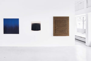 A minimalist white art gallery displaying three abstract textile artworks on a wall. From left to right: a square piece with vertical blue threads fading to black, a smaller piece with a black fringe on a white background, and a larger, brown woven hanging with a highly textured, ridged center. To the right is an opening to a brighter room with a window.