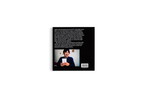 The black back cover of a book, shown against a plain white background. The cover has several paragraphs of white German text, and below it, a color photo of a smiling East Asian man in a blue top. He is holding a small, white notepad with two simple emoji faces drawn on it. A barcode is visible near the bottom.