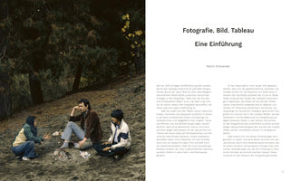 A two-page spread from a book written in German. The left page is a full-bleed color photograph of three people sitting casually on a dirt slope in a wooded area. One person gestures while speaking, a man in the center rests his head on his hand in thought, and another person sits listening. In the middle is a small pile of rocks and debris. The right page has the title Fotografie. Bild. Tableau Eine Einführung and paragraphs of text.