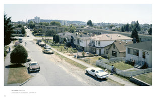 A high-angle photograph of a suburban street on a sunny day. The street slopes down, lined with modest houses and vintage cars. In the front yard of one house, two men are physically restraining and removing a third man. Other people are on the sidewalks, and in the background, an elevated railway crosses the neighborhood with a city skyline in the distance.