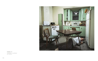 A color photograph of a man lying on the floor of a dated, mint-green kitchen. He is curled under a dark table, wide-eyed and staring forward with a look of distress, embodying the artwork's title, Insomnia.