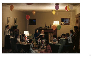 A wide, warmly lit photograph of a mid-century children's party in a living room. A woman sits in a chair holding a large, lifelike ventriloquist dummy on her lap. An audience of about ten children sits on the floor and a couch, watching the dummy intently. Colorful balloons are tied to furniture and float to the ceiling, creating a scene that is both celebratory and subtly unsettling.