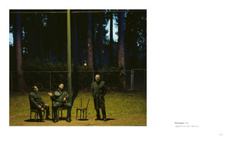 A page from a book showing a photograph titled Monologue, 2013. The photo is a cinematic, dramatically lit night scene. In a grassy area in front of a dark forest, three men are gathered under a tall pole. Two men are seated in chairs, one gesturing as if in conversation, while a third man in a long coat stands beside an empty chair.