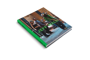 A hardcover book titled Jeff Wall, with a bright green spine, resting on a white surface. The cover features a photograph of a group of people on a city street, seen from a low, tilted angle. The title is overlaid in large, lime green text.