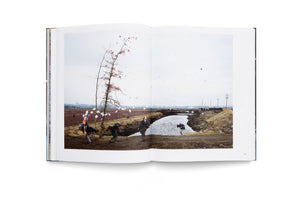 An open book with a two-page photographic spread of a chaotic outdoor scene. In a bleak, rural landscape under a white sky, several people are in motion along the muddy bank of a narrow canal. White papers fly through the air as one person does a backflip into the water.