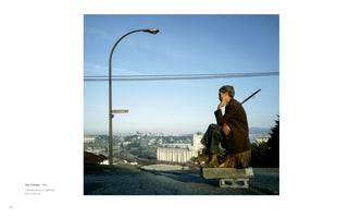 An outdoor, eye-level photograph of a man in a brown coat sitting on a tree stump that is propped up by two cinder blocks on a paved hill. He is in the pose of Rodin's The Thinker, with his chin resting on his hand, looking out over a distant city skyline under a clear blue sky. A tall, curved street lamp stands to his left.