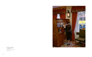 A woman with short red hair, seen from behind, stands in a richly decorated room. She wears a brown jacket and a dark skirt and is looking through a catalogue in front of a large wooden display cabinet. To her right is a window with heavy red curtains, offering a glimpse of a city street outside.