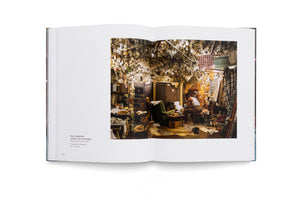 An open book displays a photograph of a man in a cluttered room. The man, seen from behind, sits on a chair under a ceiling completely covered in hundreds of glowing, bare light bulbs that brightly illuminate the space. The facing page identifies the work as After Invisible Man by Ralph Ellison, the Prologue.