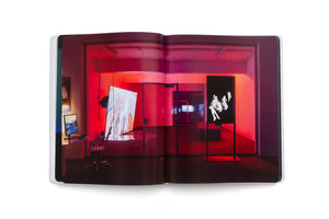 An open book displaying a photograph of a contemporary art gallery bathed in red light. The left page shows a desk and a tilted canvas with colorful abstract art. The right page features a tall black screen with a black-and-white image of floating, ghostly figures. In the background, another room with more screens is visible through a glass partition.