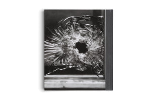 A book standing upright against a white background. The cover features a dramatic, black and white close-up photograph of a bullet hole in shattered glass, with a dark central void and cracks radiating outward. The book's dark gray, textured spine is visible on the right.
