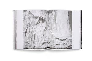 An open book with a black cover, seen from above against a white background. The pages display a full-spread, black-and-white photograph of a large, heavily textured glacier face rising out of dark, reflective water.