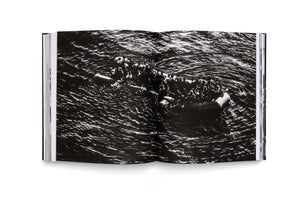 An open book on a white surface, displaying a full-bleed, black-and-white photograph across both pages. The photo, taken from a high angle, shows a large group of people crowded onto an inflatable raft in dark, choppy water.