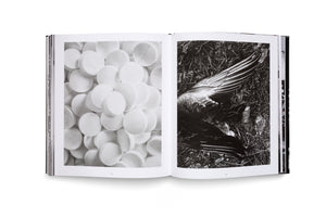 An open book on a white surface displays a two-page spread of black-and-white photographs. The photo on the left is a close-up of a pile of round, white pills. The photo on the right shows a dead bird's wing lying on dark ground covered in grass and leaves.