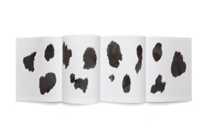 An overhead view of an accordion-folded book open on a white background. The white pages feature several irregularly shaped black spots with a furry texture, creating a pattern that resembles cowhide.