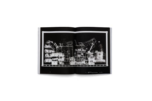 A high-angle photograph of an open book lying on a white surface. The book displays a two-page spread featuring a black-and-white X-ray image. The radiograph shows a dense, chaotic assembly of various objects, including musical instruments and household items, crowded onto shelves. The objects appear as ghostly, translucent white outlines against the black background, overlapping to create a complex abstract composition.