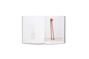 An open art book on a white background. The left page shows a watercolor drawing of a person standing with two crutches shaped like long, reddish-brown bones. The right page displays a photograph of two similar, sculptural bone-like crutches leaning against a white wall in a corner.