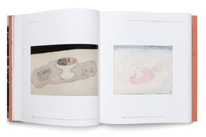 An open art book displays two paintings on a two-page spread. The left page shows a still life with a fruit bowl, basket, and cup on a table. The right page features a simple painting of a pink cat lying on its back, playing with a butterfly against a white background. Each artwork is accompanied by a caption.