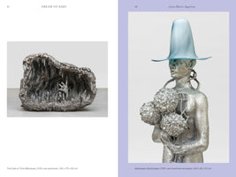 An open art book displaying two sculptures on facing pages. On the left, a cast aluminum sculpture shows a rocky grotto with a single hand reaching out from the dark interior. On the right, a cast aluminum sculpture of a person holds a bouquet of metallic hydrangeas and wears a large, translucent blue glass hat.