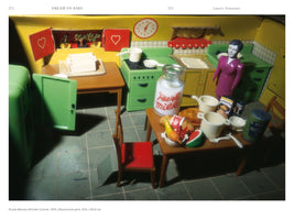 A photograph from a book showing Laurie Simmons's artwork Purple Woman/Kitchen/Corner, 1979. The image is a close-up of a vintage dollhouse kitchen with yellow walls, bright green plastic appliances, and a tiled floor. A female doll in a purple dress stands by a counter. In the foreground, a table is set with miniature food, including a large glass bottle of milk, bread, fruit, and a can of baked beans. Harsh lighting casts strong shadows across the scene.