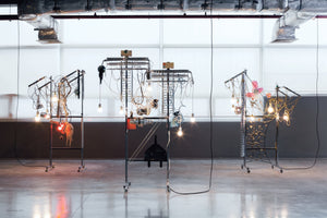 An art installation of three complex, machine-like sculptures on rolling metal frames stands in a spacious gallery with a concrete floor. Each sculpture is a chaotic assembly of wires, glowing lightbulbs, and various found objects. Exposed silver ductwork hangs from the ceiling above the display.