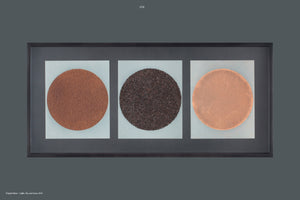 A contemporary art piece in a long, horizontal black frame against a dark gray background. The triptych displays three distinct circles, each centered on a light-colored square. From left to right, there is a circle of fine, reddish-brown granules; a circle of coarse, dark brown granules; and a smooth, solid, coppery-brown disc. The artwork uses coffee, tea, and cacao.