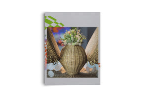 A light gray booklet presented on a white background. The cover features a central photograph of a decorative, light brown woven wicker vase with two wing-like sides. The vase is topped with a small bouquet of green succulents and white flowers, and decorated with dangling, iridescent shell discs. In the upper left corner of the cover, there is a green and white geometric pattern.