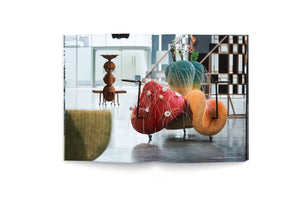 An open book displays a photograph of an art installation in a modern gallery. The central piece is a large, abstract sculpture on wheels, composed of colorful, woven, organic shapes. The forms are red, orange, and a gradient of green and yellow. The red section is adorned with thin white tendrils. In the background, another wooden sculpture and a geometric screen are visible.