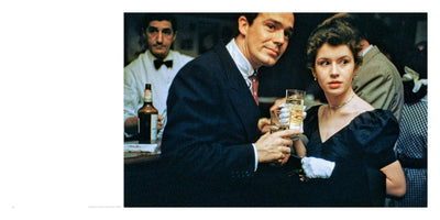 A vintage color photo from 1949 of a couple at a party in New York. A man in a dark suit and tie holds a glass and smiles at the camera, while a woman in a black puff-sleeve dress and white gloves holds her glass and looks off to the side. A bartender stands behind them at the bar.