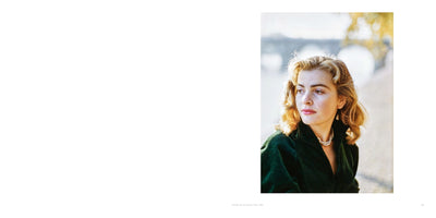A vintage color photograph of a young woman with wavy blonde hair and a pensive expression, looking to her left. She is wearing an elegant dark green corduroy jacket and a double-strand pearl necklace. The outdoor setting is softly focused, showing a bridge and autumn trees in warm sunlight.