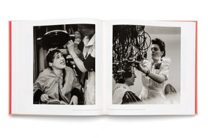 An open book with a red spine lays flat, displaying a two-page spread of black and white photographs from a 1940s beauty parlor. On the left, a woman smiles while sitting under a large dome hair dryer. On the right, another woman receives a perm from a complex machine with wires and curlers as a stylist makes adjustments.